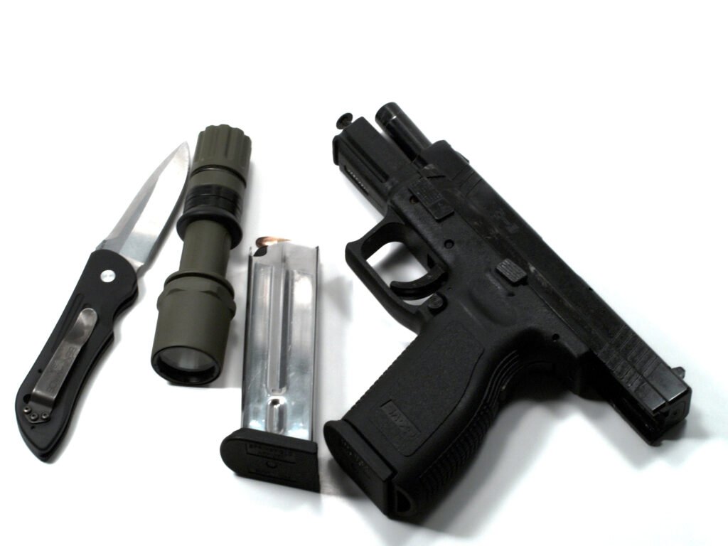 A black handgun, metal magazine, folding knife, and green flashlight are arranged on a white background. The items suggest a tactical or security context.